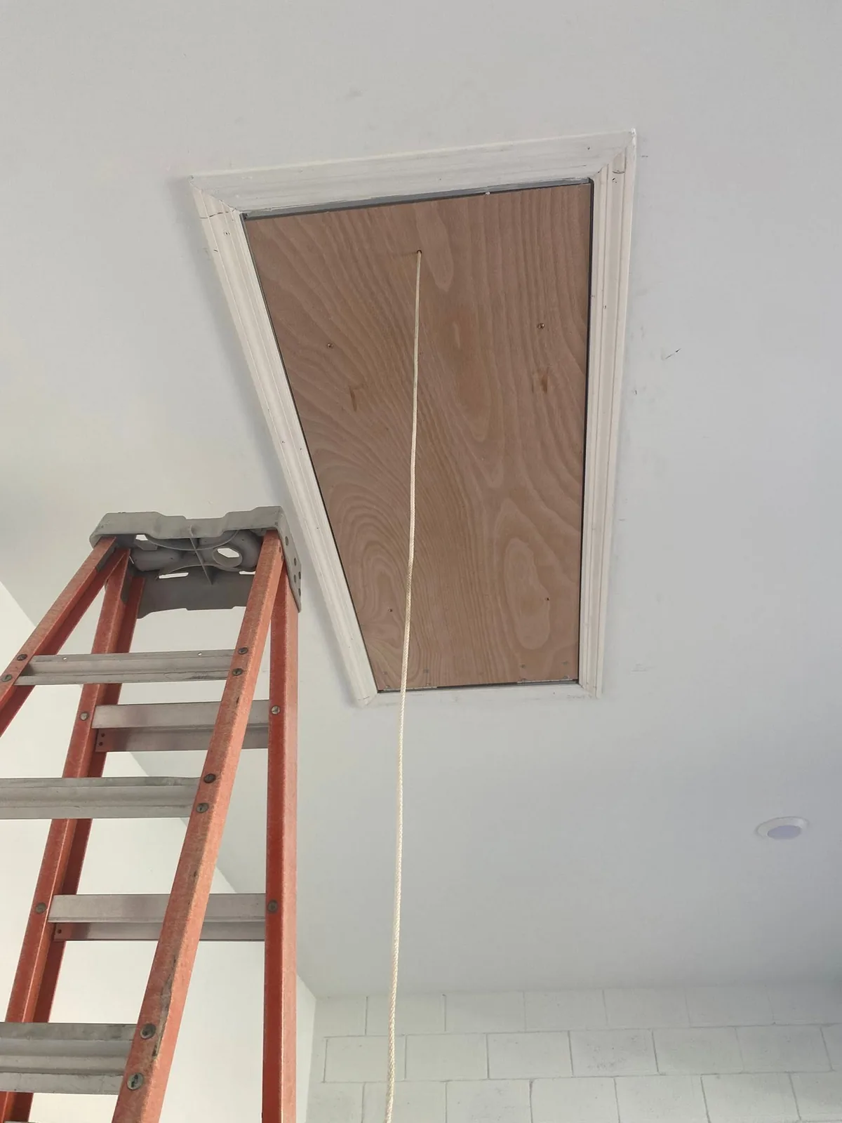 Panel de madera de acceso al techo con moldura blanca y cordón, con una escalera debajo.